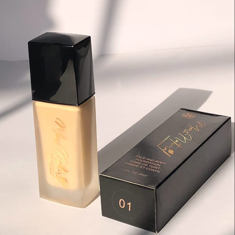 A bottle of liquid foundation with a black cap stands next to its black retail box on a white surface. The foundation liquid is a warm light beige, and the packaging features gold foil branding and the shade number 01.