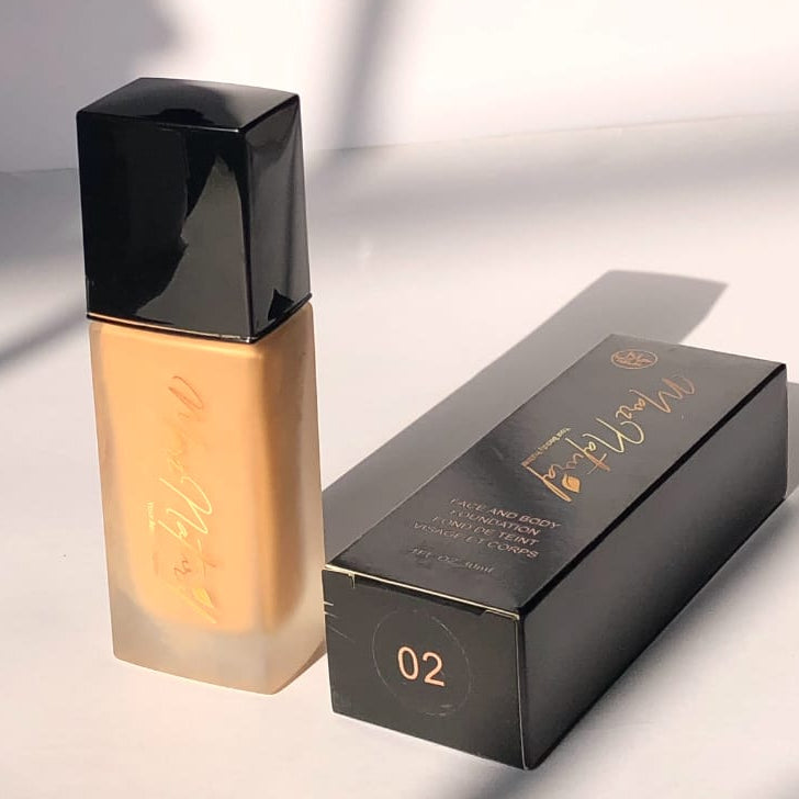 A bottle of liquid foundation with a black cap stands next to its black retail box on a white surface. The foundation liquid is a warm tan shade, and the box has gold branding and the shade number 02.