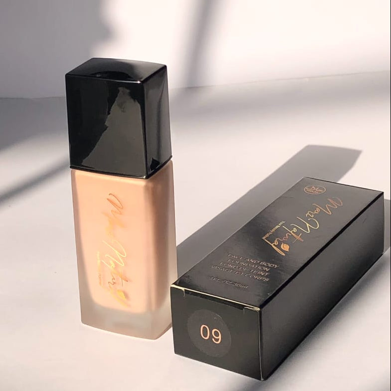 A bottle of liquid foundation with a frosted beige body and a glossy black cap stands next to its black retail box on a white surface. The box has gold script branding and the shade number 09 printed in gold on the bottom.