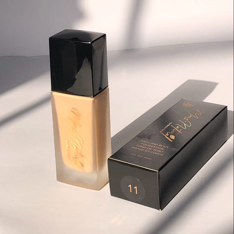 A bottle of liquid foundation with a black cap stands next to its black retail box on a white surface. The foundation bottle contains a warm beige liquid and has gold script branding.