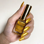 A hand with long, metallic gold manicured nails holds a bottle of Nailah Helly Shimmer Gold Bubble Nail Polish against a plain off-white background.