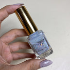 A hand holding a bottle of Morphe Nails 'Bling Out' glitter nail polish. The nail polish is a light lavender-blue color with dense silver, pink, and blue glitter, and is housed in a clear glass bottle with a shiny gold cap.