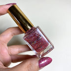 A hand holding a bottle of glitter nail polish. The nail polish inside the clear glass bottle is a sparkling magenta-pink color with holographic glitter.