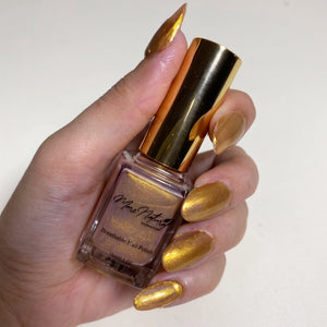 A hand holding a bottle of gold metallic nail polish against a plain light background. The nails are painted in a matching shimmering gold shade, and the bottle has a shiny gold cap.