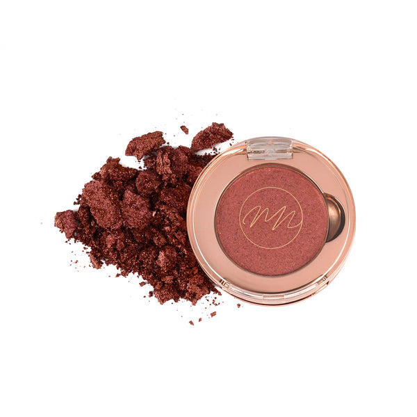 A pan of copper brown eyeshadow with loose powder scattered outside the pan, on a white background.