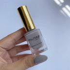 A hand holding a bottle of Max Matar Breathable Nail Polish with a metallic gold cap, showing a light gray nail polish inside the clear glass bottle.