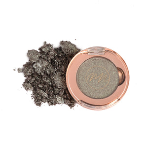 A pan of chromatic mirage eye shadow with a light green shade, alongside loose eye shadow powder scattered outside the pan.