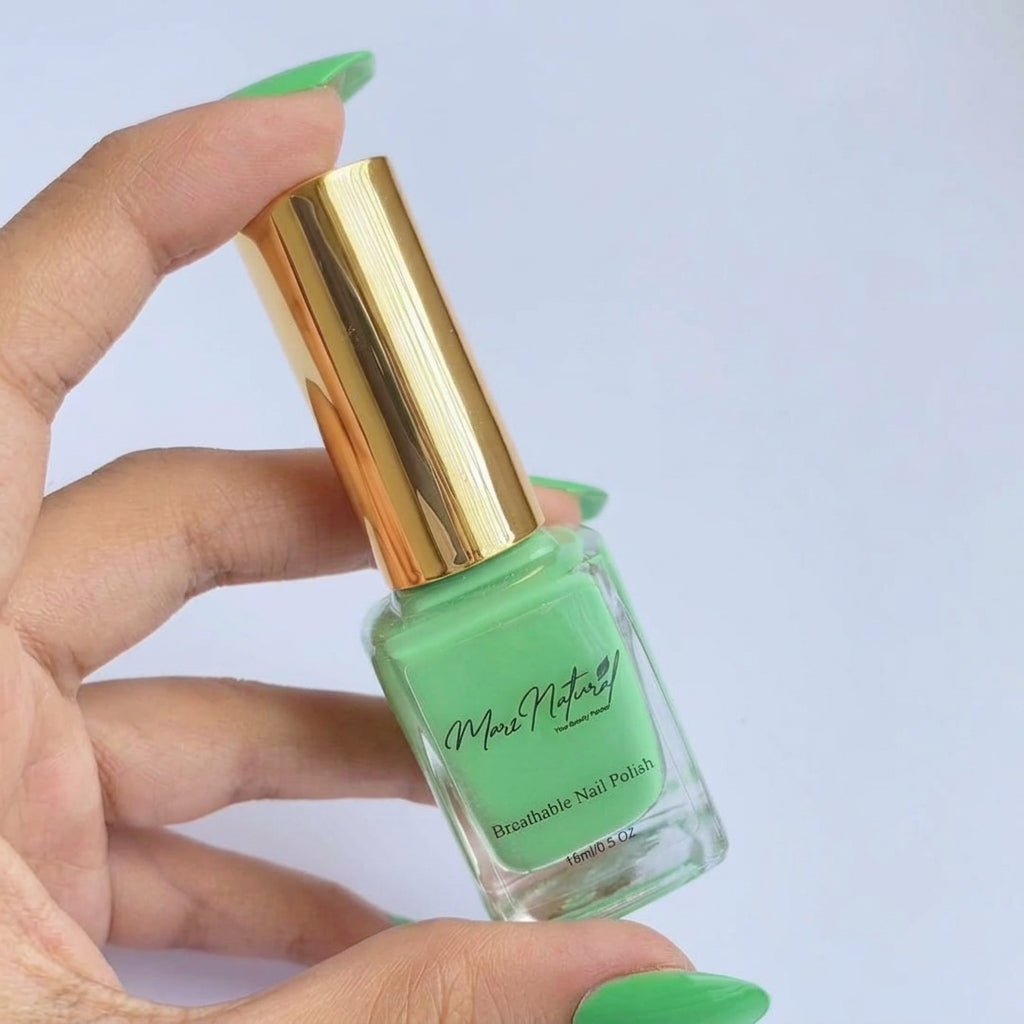 A hand with mint green nails holds a bottle of mint green breathable nail polish with a shiny gold cap against a light blue background.
